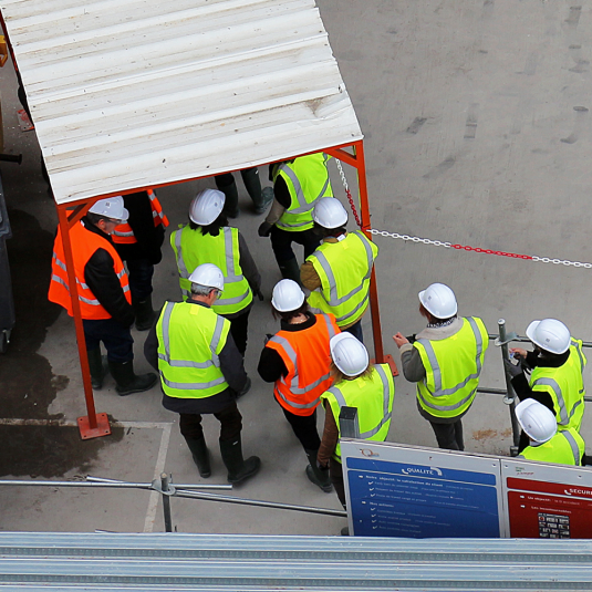 Construction workers with yellow and orange high-visibility vests and white helmets enter the construction site.