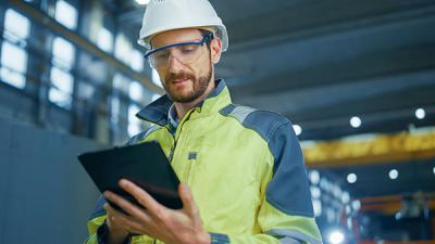 The digital supply chain: Man on construction site with helmet and yellow high-visibility waistcoat holding a tablet.