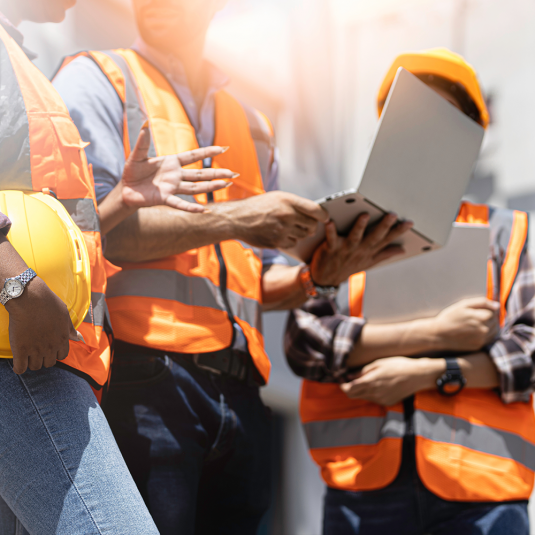Three workers at the construction site are talking to each other, lookin.g at a laptop.